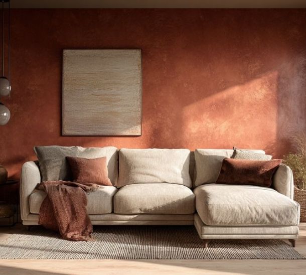 Soggiorno moderno e accogliente con divano beige a penisola, parete color terracotta con finitura materica, cuscini e plaid nei toni caldi, tappeto chiaro, tavolino in ceramica e luce naturale che entra da una grande finestra laterale.